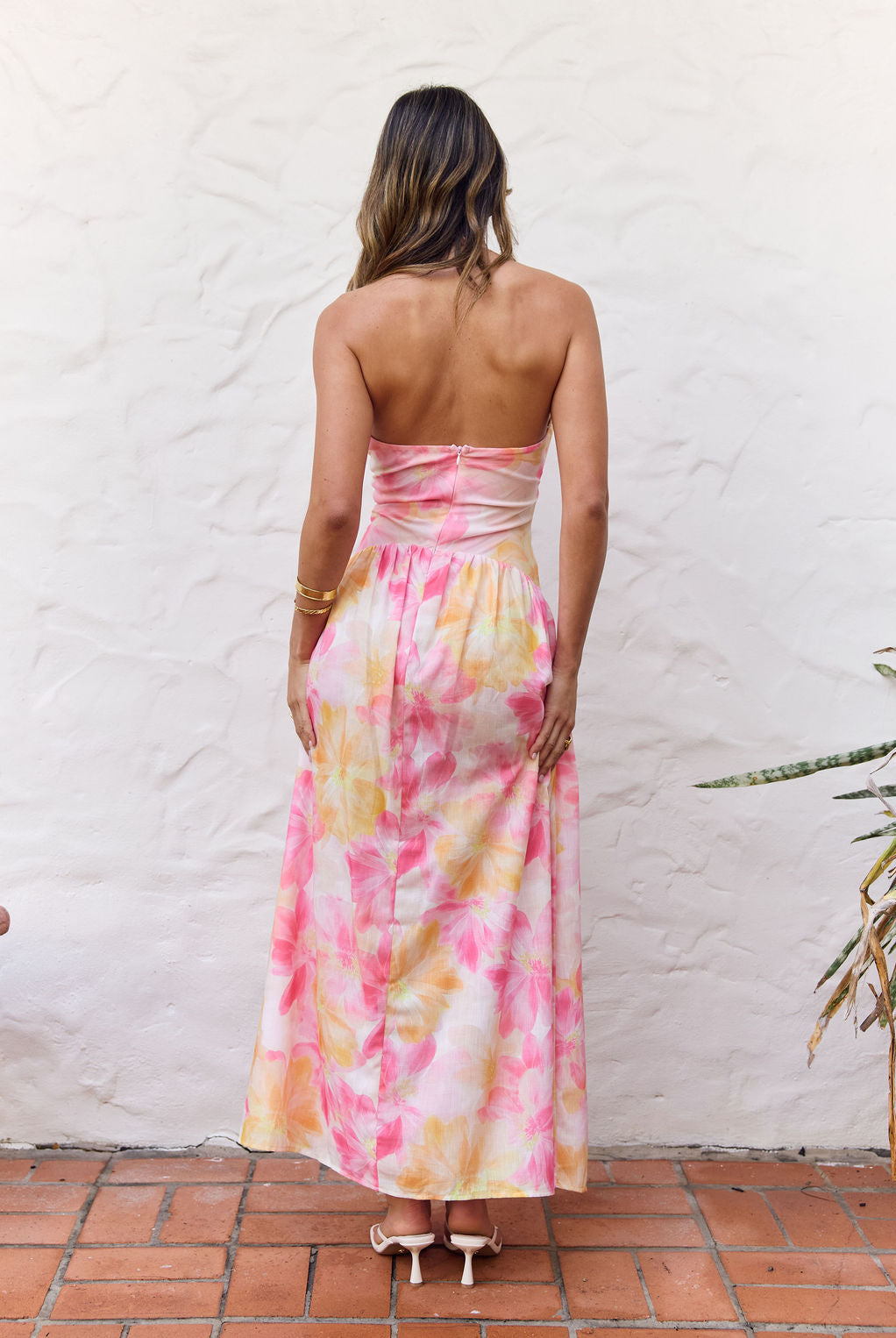 A woman viewed from behind wearing a strapless maxi dress with a tie-dye pattern in pink, yellow, and orange on a white base, standing against a white stucco wall.