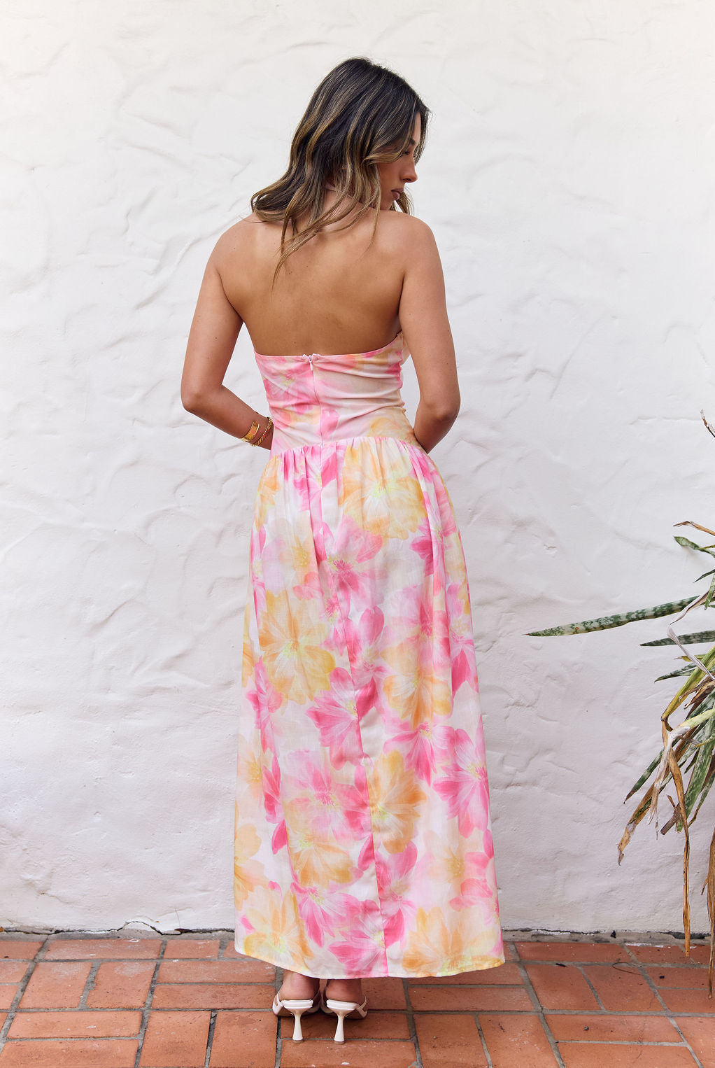 A woman viewed from behind wearing a strapless maxi dress with a pink, yellow, and white tie-dye pattern, standing against a white stucco wall.