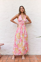 A woman modeling a pink, yellow, and white watercolor floral halter maxi dress, standing against a white stucco wall on a terracotta tile floor.