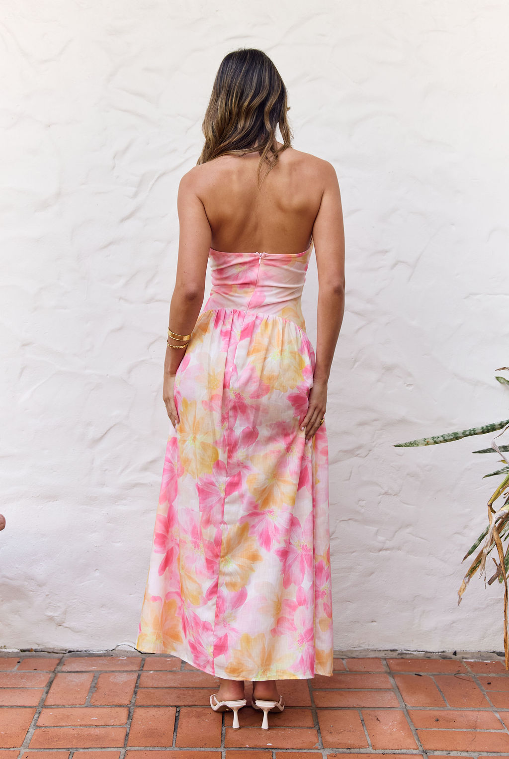 A woman viewed from behind wearing a strapless maxi dress with a tie-dye pattern in pink, yellow, and orange on a white base, standing against a white stucco wall.