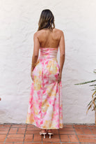 A woman viewed from behind wearing a strapless maxi dress with a tie-dye pattern in pink, yellow, and orange on a white base, standing against a white stucco wall.