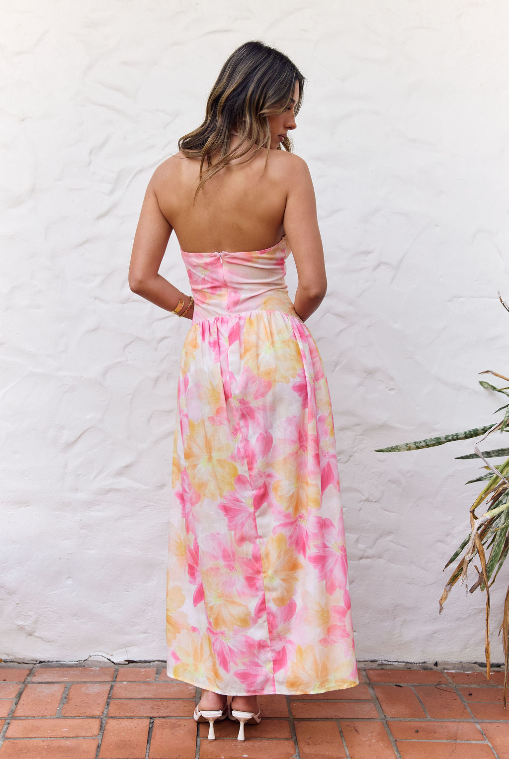 A woman viewed from behind wearing a strapless maxi dress with a pink, yellow, and white tie-dye pattern, standing against a white stucco wall.